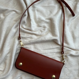 Puede incluir: Bolso bandolera de cuero marrón con una correa larga y ajustable. El bolso tiene forma rectangular con esquinas redondeadas y se asegura con dos tachuelas doradas. La correa se sujeta con cierres dorados.