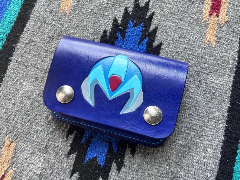 May include: A blue leather wallet with a stylized, light blue and red emblem on the front. The wallet has two silver snap closures and is likely designed to hold cards and cash. The item is placed on a patterned textile.