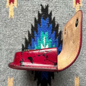 May include: A red leather case with black paint splatters, open to reveal a tan interior. The case has a curved shape and is secured with metal snaps. The item is likely a small accessory holder.