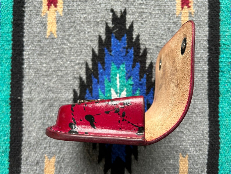 May include: A red leather case with black paint splatters, open to reveal a tan interior. The case has a curved shape and is secured with metal snaps. The item is likely a small accessory holder.