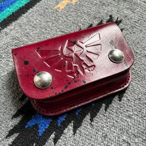May include: A burgundy leather case with a raised design and silver button closures. The case features a splattered paint effect and a stylized emblem. The case is rectangular and appears to be handmade.