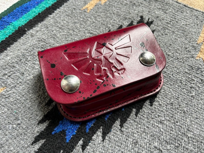 May include: A burgundy leather case with a raised design and silver button closures. The case features a splattered paint effect and a stylized emblem. The case is rectangular and appears to be handmade.