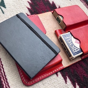Può includere: Un taccuino nero con un elastico è appoggiato su un porta carte in pelle rossa. Il porta carte contiene due mazzi di carte. Lo sfondo è un tessuto fantasia.