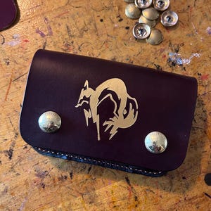 May include: A burgundy leather wallet with a gold fox design and two brass snap closures. The wallet is rectangular and closed, with a stitched edge. Several brass snaps are scattered nearby on a wooden surface.