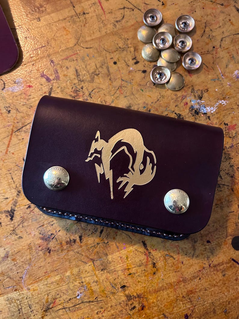 May include: A burgundy leather wallet with a gold fox design and two brass snap closures. The wallet is rectangular and closed, with a stitched edge. Several brass snaps are scattered nearby on a wooden surface.