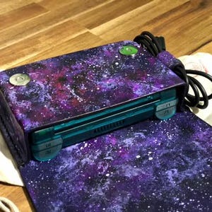 May include: A purple and blue galaxy-themed crossbody bag with a teal-colored handheld gaming device inside. The bag has a magnetic closure and a black cord. The bag is open, revealing the interior.