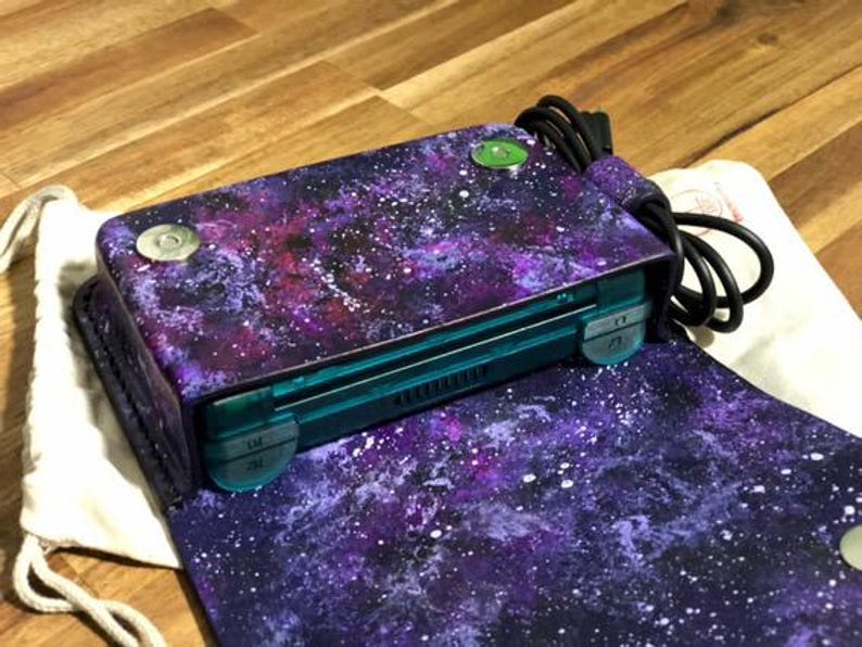 May include: A purple and blue galaxy-themed crossbody bag with a teal-colored handheld gaming device inside. The bag has a magnetic closure and a black cord. The bag is open, revealing the interior.