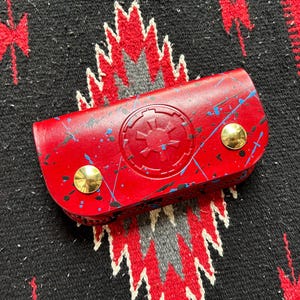 Pode incluir: Um estojo de couro vermelho com um design de respingos pintados em azul e preto. O estojo apresenta um emblema em relevo e é fixado com dois rebites dourados. O estojo está sobre um tecido estampado em preto e vermelho.