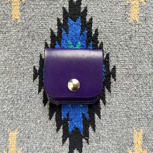 Puede incluir: Una peque&ntilde;a bolsa de cuero p&uacute;rpura con un cierre de bot&oacute;n a presi&oacute;n dorado. La bolsa est&aacute; colocada sobre un fondo textil con estampado gris, azul y negro.