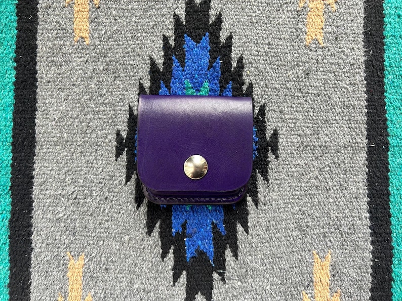 Puede incluir: Una peque&ntilde;a bolsa de cuero p&uacute;rpura con un cierre de bot&oacute;n a presi&oacute;n dorado. La bolsa est&aacute; colocada sobre un fondo textil con estampado gris, azul y negro.