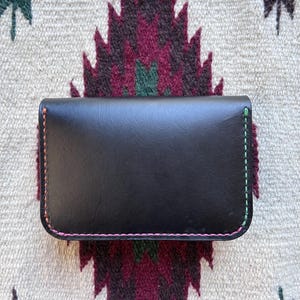 May include: A black leather wallet with colorful stitching. The wallet is rectangular with rounded corners and features pink, green, and orange stitching along the edges. The wallet is resting on a patterned textile with a geometric design.