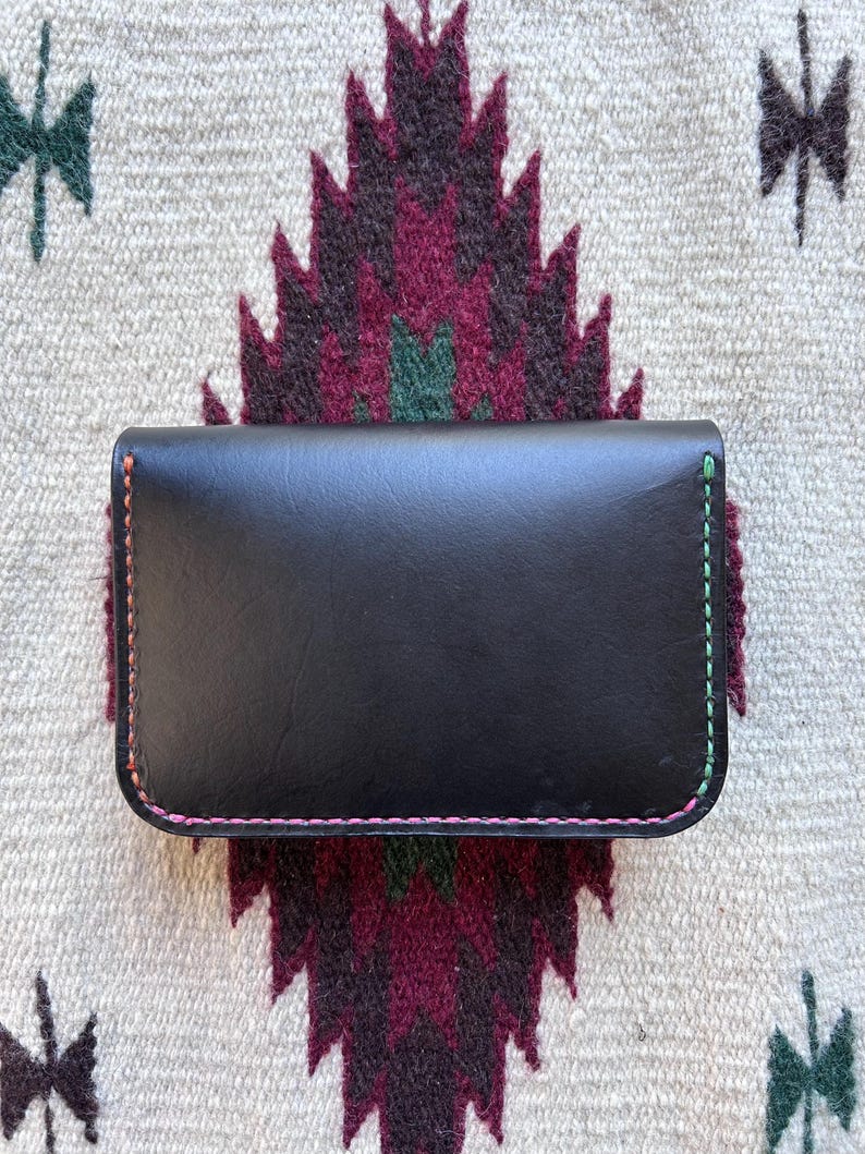 May include: A black leather wallet with colorful stitching. The wallet is rectangular with rounded corners and features pink, green, and orange stitching along the edges. The wallet is resting on a patterned textile with a geometric design.