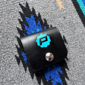 Puede incluir: Cartera de cuero negro con un logotipo "P" turquesa en un dise&ntilde;o en forma de nube. La cartera tiene un cierre de bot&oacute;n a presi&oacute;n plateado y est&aacute; sobre un fondo textil estampado gris y azul.