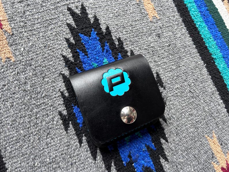 Puede incluir: Cartera de cuero negro con un logotipo "P" turquesa en un dise&ntilde;o en forma de nube. La cartera tiene un cierre de bot&oacute;n a presi&oacute;n plateado y est&aacute; sobre un fondo textil estampado gris y azul.