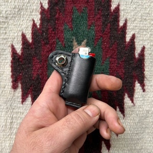 Puede incluir: Una funda para encendedor de cuero negro con un anillo de metal plateado. La funda está diseñada para contener un encendedor de tamaño estándar.