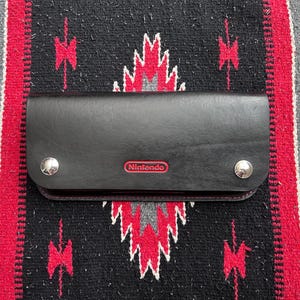 以下が含まれることがあります： 赤色のNintendoロゴが入った黒い革製のNintendo Switchケース。ケースには2つのシルバーのスナップ留めと赤いステッチの縁取りがあります。ケースは、黒、赤、白の模様のテキスタイル上にあります。