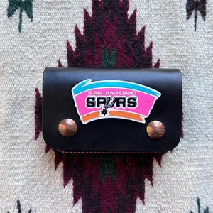 May include: Black leather wallet featuring the San Antonio Spurs logo in pink, teal, and orange. The wallet has two copper-colored snap closures. The background is a patterned textile.