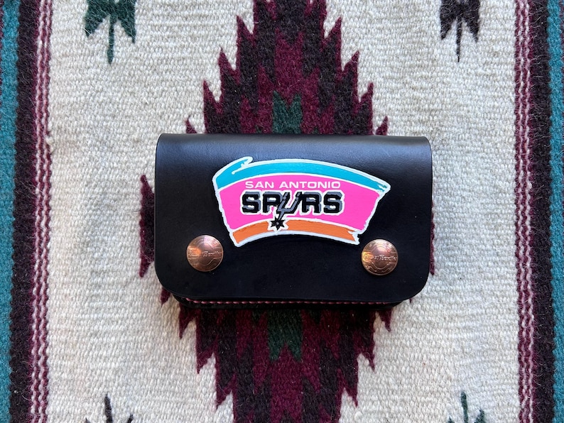 May include: Black leather wallet featuring the San Antonio Spurs logo in pink, teal, and orange. The wallet has two copper-colored snap closures. The background is a patterned textile.