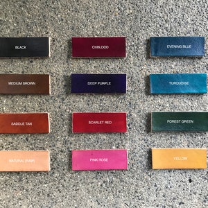 May include: A collection of rectangular leather swatches in various colors, including black, oxblood, evening blue, medium brown, deep purple, turquoise, saddle tan, scarlet red, forest green, natural (raw), pink rose, and yellow. Each swatch has the color name printed on it.