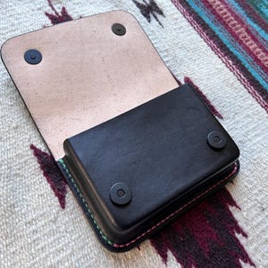 May include: Black leather wallet with a flap, open to reveal a light tan interior. The wallet has a rectangular shape with rounded corners and is secured with two black snap closures. The edges are stitched with pink and green thread.