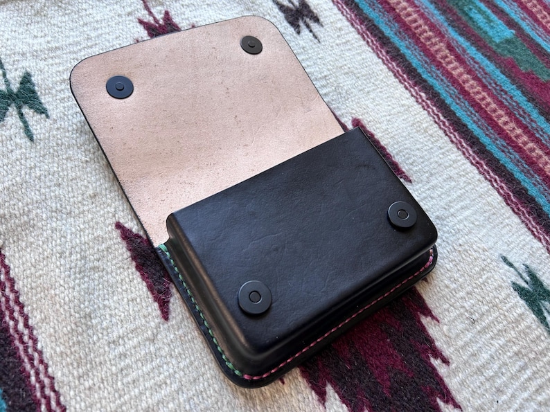 May include: Black leather wallet with a flap, open to reveal a light tan interior. The wallet has a rectangular shape with rounded corners and is secured with two black snap closures. The edges are stitched with pink and green thread.