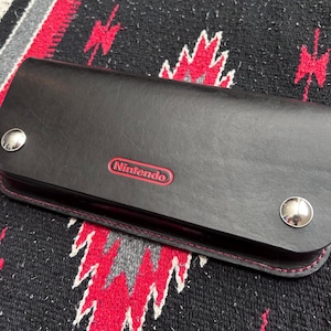 May include: Black leather wallet with the red "Nintendo" logo. The wallet has a snap closure and red stitching. The wallet is rectangular and appears to be made of high-quality leather. The background is a patterned textile.