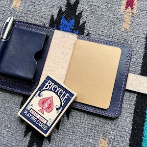 Può includere: Una copertina per taccuino in pelle blu navy con penna, porta carte e un piccolo blocco note. Un mazzo di carte Bicycle è appoggiato accanto al taccuino aperto. La copertina è cucita con filo rosa.