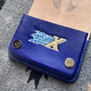 May include: A blue leather wallet with the Mega Man X logo in silver and gold. The wallet has a snap closure and blue stitching. The interior is a light tan color. The wallet is on a gray patterned surface.