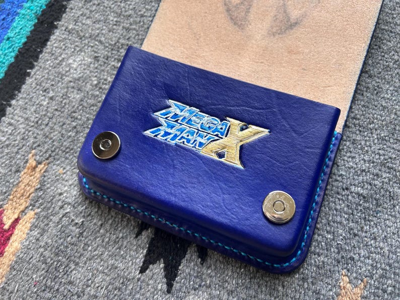 May include: A blue leather wallet with the Mega Man X logo in silver and gold. The wallet has a snap closure and blue stitching. The interior is a light tan color. The wallet is on a gray patterned surface.
