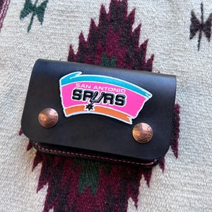 May include: Black leather wallet featuring the San Antonio Spurs logo in pink, teal, and orange. The wallet has two copper-colored snap closures. The background is a patterned textile.