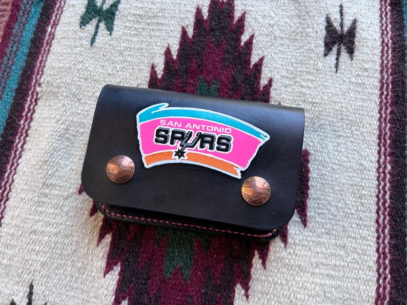 May include: Black leather wallet featuring the San Antonio Spurs logo in pink, teal, and orange. The wallet has two copper-colored snap closures. The background is a patterned textile.