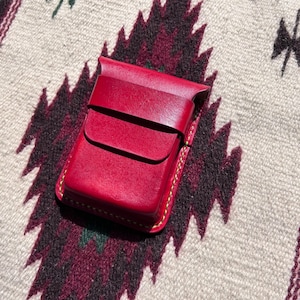 Könnte beinhalten: Ein rotes Lederetui mit Klappenverschluss. Das Etui ist rechteckig mit abgerundeten Ecken und gelben Nähten. Das Etui liegt auf einem gemusterten Textil.