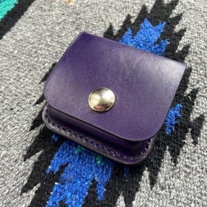 Può includere: Un piccolo astuccio in pelle viola con patta arrotondata e chiusura a scatto color oro. L'astuccio è appoggiato su un tessuto a fantasia grigio e blu.