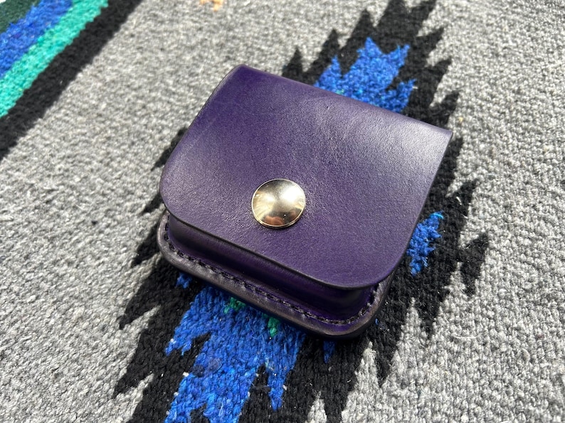 Puede incluir: Una peque&ntilde;a bolsa de cuero p&uacute;rpura con una solapa redondeada y un cierre de bot&oacute;n a presi&oacute;n dorado. La bolsa est&aacute; sobre un textil estampado en gris y azul.