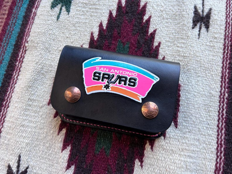 May include: Black leather wallet featuring the San Antonio Spurs logo in pink, teal, and orange. The wallet has two copper-colored snap closures and a stitched edge. The wallet is displayed on a patterned textile.