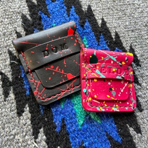 May include: Two small leather wallets with a splatter paint design. One wallet is black with red paint and the name "DOM" embossed on the front. The other wallet is pink with multi-colored paint and the name "REIDDE" embossed on the front.
