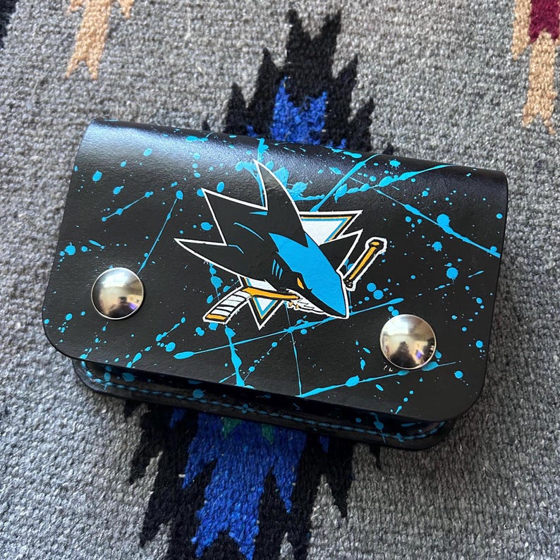 May include: Black leather wallet featuring the San Jose Sharks logo with a teal and white design. The wallet has a splatter paint effect in teal and two silver snap closures. The wallet is closed.