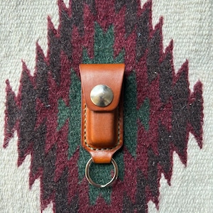 Puede incluir: Un llavero de cuero marrón con un anillo de metal plateado y un cierre a presión. El llavero está sobre una alfombra estampada.