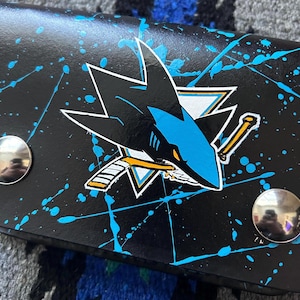 May include: Black wallet with the San Jose Sharks logo in teal, white, and black. The wallet has a teal splatter paint design and two silver snap closures. The Sharks logo is a stylized shark head with a hockey stick.