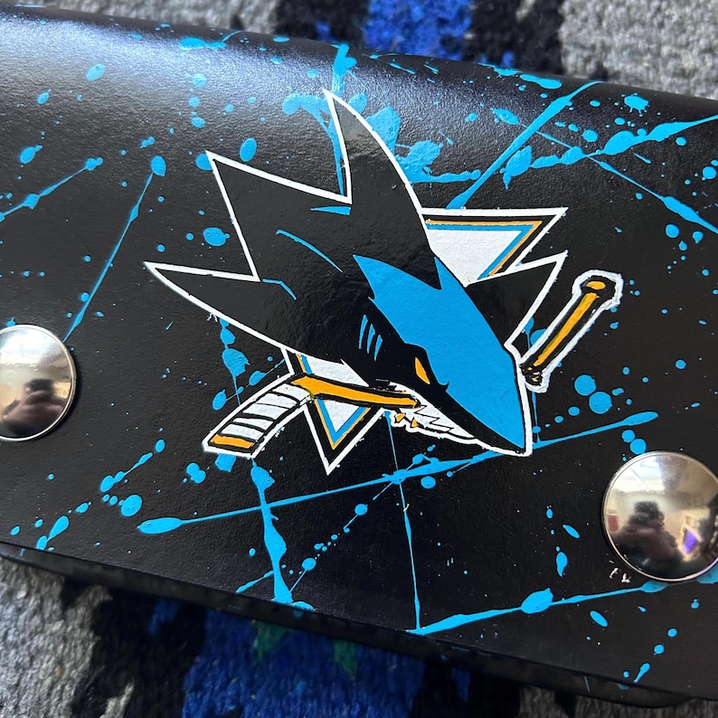 May include: Black wallet with the San Jose Sharks logo in teal, white, and black. The wallet has a teal splatter paint design and two silver snap closures. The Sharks logo is a stylized shark head with a hockey stick.