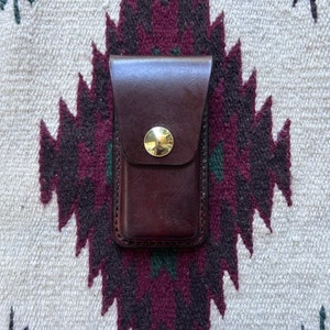 以下が含まれることがあります： 真鍮製のボタン留め具が付いた、茶色の革製のポケットナイフケースです。