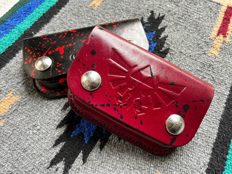 May include: Two leather pouches, one black with red splatter and one red with black splatter, are displayed. The red pouch features an embossed design. Both have silver snap closures. The pouches are on a patterned textile.