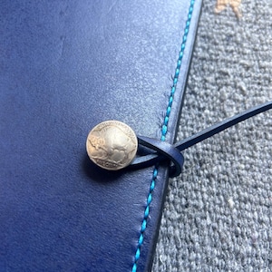 May include: A dark blue leather notebook with a blue stitching detail and a silver coin closure. The notebook is lying on a gray textured surface.