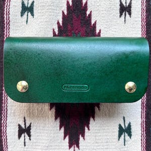 May include: A green leather case with gold accents and the word "Nintendo" embossed on the front.