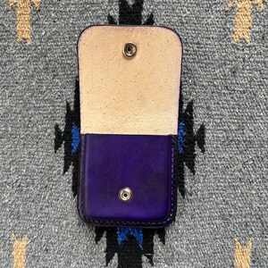 Puede incluir: Peque&ntilde;o estuche de cuero morado abierto, con interior de color natural. El estuche es rectangular con esquinas redondeadas y cierre de bot&oacute;n a presi&oacute;n. El fondo es un textil gris estampado.