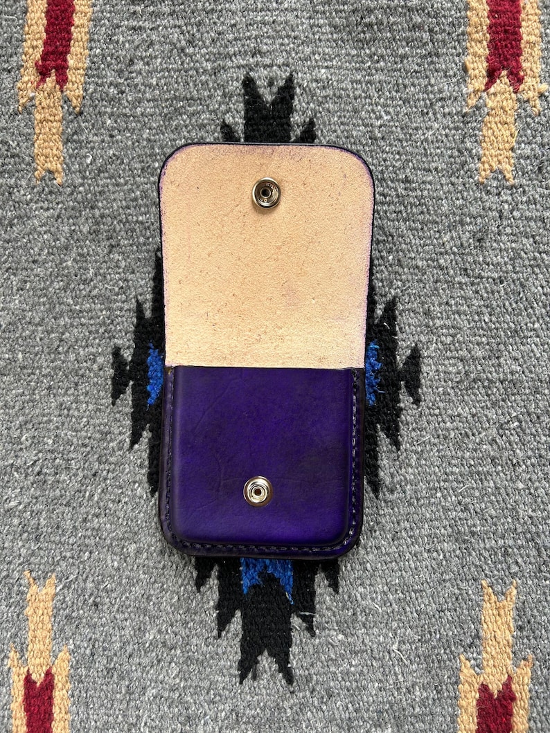 Puede incluir: Peque&ntilde;o estuche de cuero morado abierto, con interior de color natural. El estuche es rectangular con esquinas redondeadas y cierre de bot&oacute;n a presi&oacute;n. El fondo es un textil gris estampado.