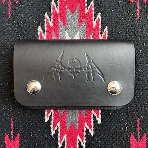 May include: Black leather wallet with a decorative embossed design featuring a skull and wing motif. The wallet is rectangular with rounded edges and secured by two silver snap closures. The wallet is displayed on a patterned textile.