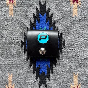 Puede incluir: Peque&ntilde;o estuche de cuero negro con cierre de bot&oacute;n a presi&oacute;n plateado. Presenta una letra "P" turquesa dentro de un dise&ntilde;o floral estilizado. El fondo es un textil gris con un patr&oacute;n geom&eacute;trico.