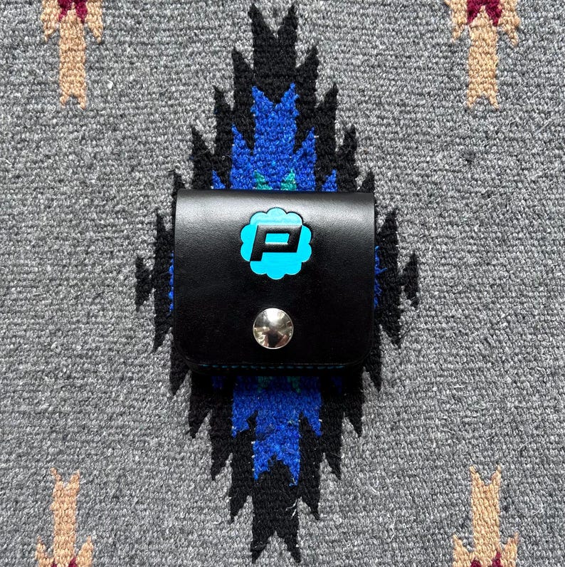 Puede incluir: Peque&ntilde;o estuche de cuero negro con cierre de bot&oacute;n a presi&oacute;n plateado. Presenta una letra "P" turquesa dentro de un dise&ntilde;o floral estilizado. El fondo es un textil gris con un patr&oacute;n geom&eacute;trico.