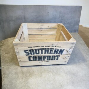 Pode incluir: Uma caixa de madeira com as palavras "SOUTHERN COMFORT" em letras pretas. O texto "THE SPIRIT OF NEW ORLEANS" e "EST. 1874" tamb&eacute;m s&atilde;o vis&iacute;veis. A caixa &eacute; feita de madeira clara e est&aacute; sobre uma superf&iacute;cie cinza.